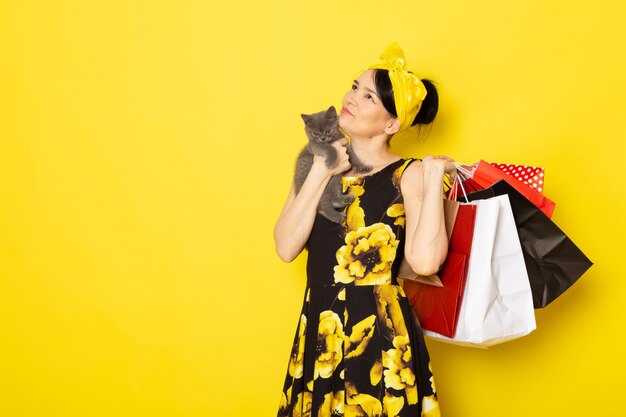 Une vue de face jeune femme en fleur jaune-noir robe conçue avec un bandage jaune sur la tête tenant des colis et chaton sur le jaune