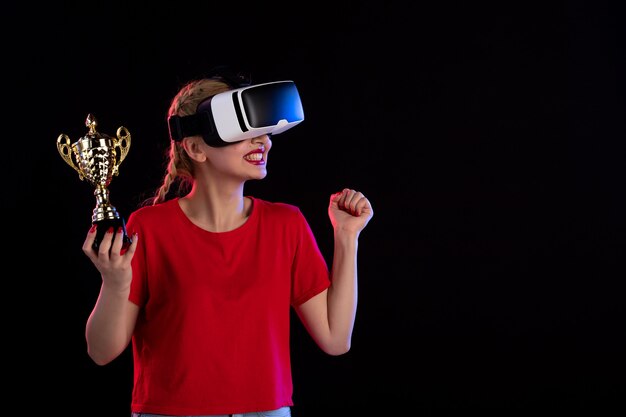 Vue de face d'une jeune femme jouant à la réalité virtuelle et gagnant la coupe sur un mur sombre