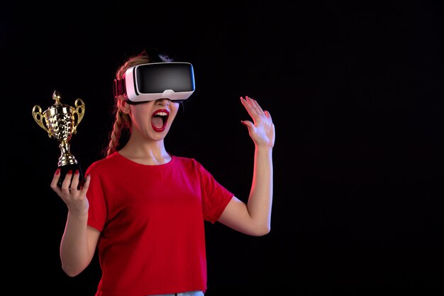 Vue de face d'une jeune femme jouant à la réalité virtuelle et gagnant la coupe sur un mur sombre