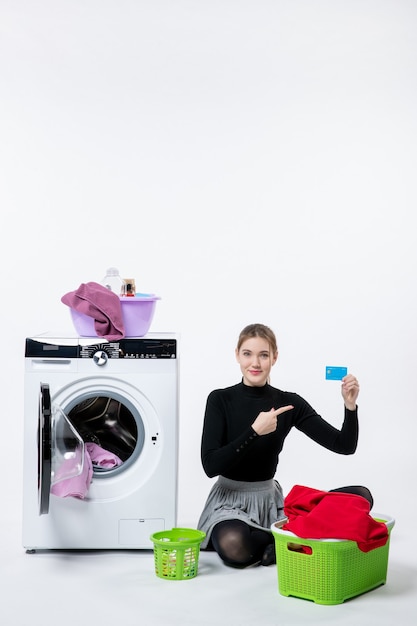 Vue de face d'une jeune femme avec une machine à laver tenant une carte bancaire sur un mur blanc