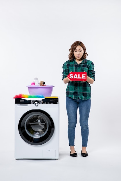 Vue de face d'une jeune femme avec une machine à laver tenant une vente par écrit sur le mur blanc