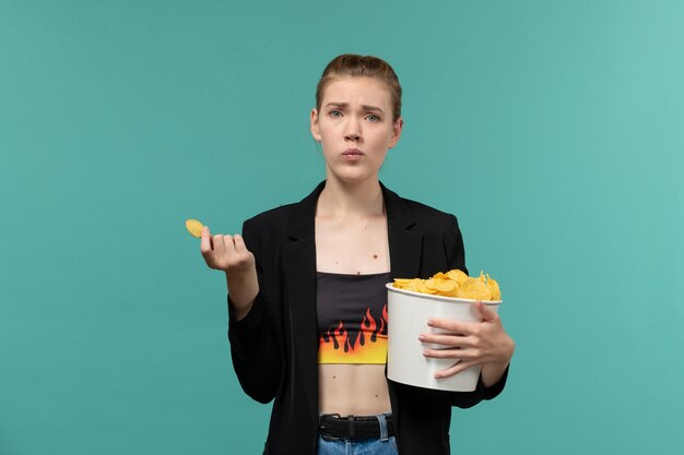 Vue de face jeune femme mangeant des chips de pomme de terre regarder un film sur la surface bleu clair
