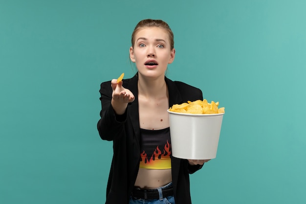 Vue de face jeune femme mangeant des chips de pomme de terre regarder un film sur la surface bleue
