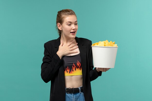 Vue de face jeune femme mangeant des chips de pomme de terre regarder un film sur la surface bleue