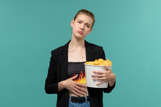 Vue de face jeune femme mangeant des cips de pommes de terre et regarder un film sur une surface bleue