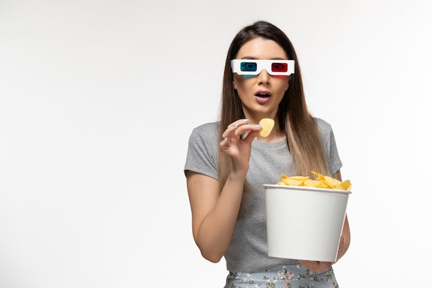 Vue de face jeune femme mangeant des cips tout en regardant un film dans des lunettes de soleil sur une surface blanche légère