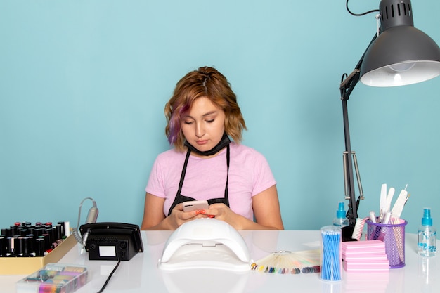 Une vue de face jeune femme manucure en t-shirt rose avec des gants noirs et un masque noir assis devant la table et à l'aide d'un téléphone sur bleu