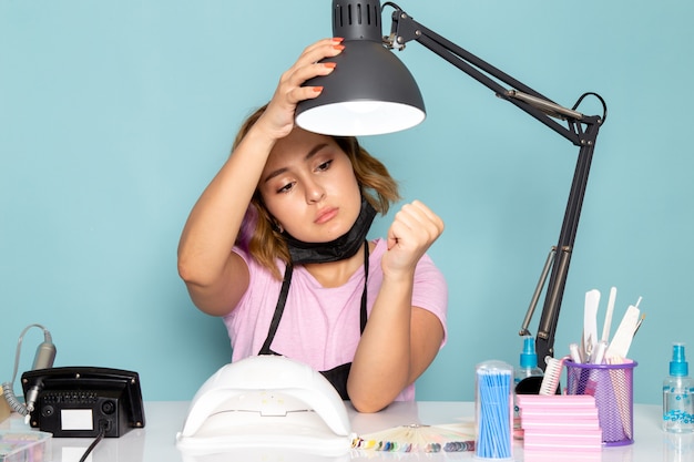 Une vue de face jeune femme manucure en t-shirt rose avec des gants noirs et un masque noir assis devant la table sur le bleu