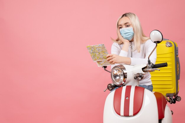 Vue de face jeune femme avec masque sur cyclomoteur avec valise jaune en regardant la carte