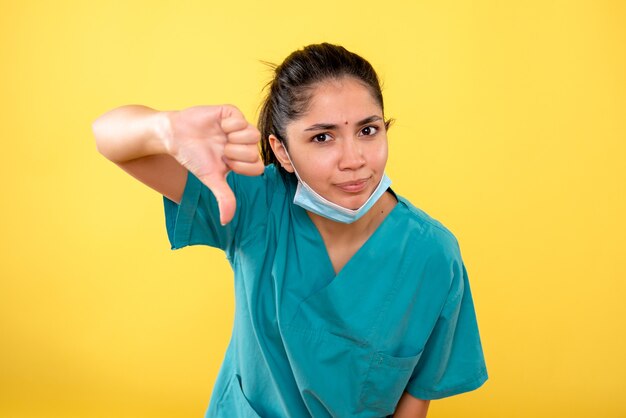 Vue de face de la jeune femme avec masque médical faisant thum down signe sur mur isolé jaune