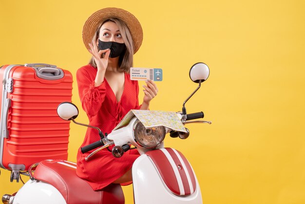 Vue de face jeune femme avec masque noir sur un cyclomoteur tenant un billet faisant un signe savoureux