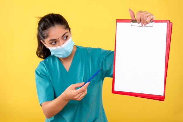 Vue de face de la jeune femme avec masque tenant le presse-papiers sur le mur jaune