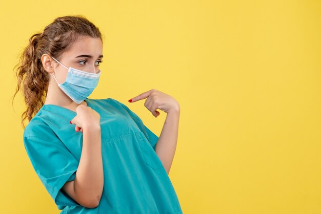 Vue de face de la jeune femme médecin en chemise médicale et masque stérile sur mur jaune