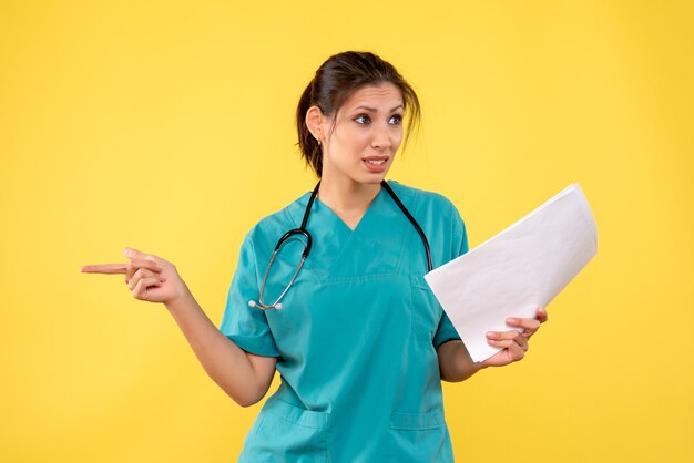 Vue de face jeune femme médecin en chemise médicale tenant l'analyse de papier sur fond jaune