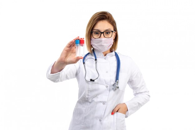 Une vue de face jeune femme médecin en combinaison médicale blanche avec stéthoscope portant un masque de protection blanc tenant des flacons sur le blanc