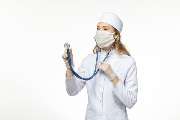 Vue de face jeune femme médecin en combinaison médicale portant un masque de protection en raison d'un coronavirus à l'aide d'un stéthoscope sur une surface blanche