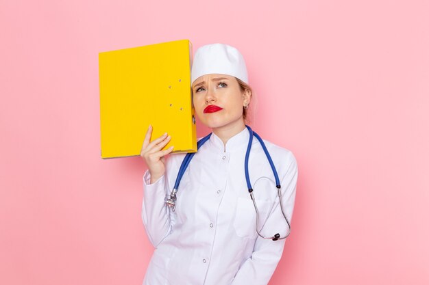 Vue de face jeune femme médecin en costume blanc avec stéthoscope bleu tenant des fichiers jaunes sur le travail de fille de l'espace rose