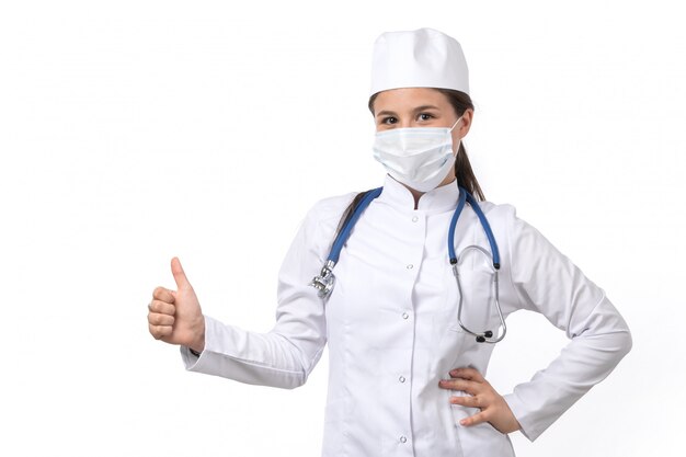 Une vue de face jeune femme médecin en costume médical blanc et bonnet blanc avec stéthoscope bleu portant un masque