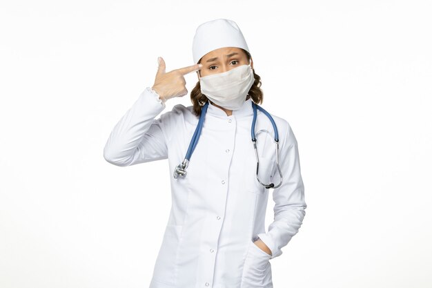 Vue de face jeune femme médecin en costume médical blanc et avec masque en raison d'un coronavirus sur le sol blanc