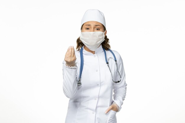 Vue de face jeune femme médecin en costume médical blanc et avec masque en raison d'un coronavirus sur surface blanche