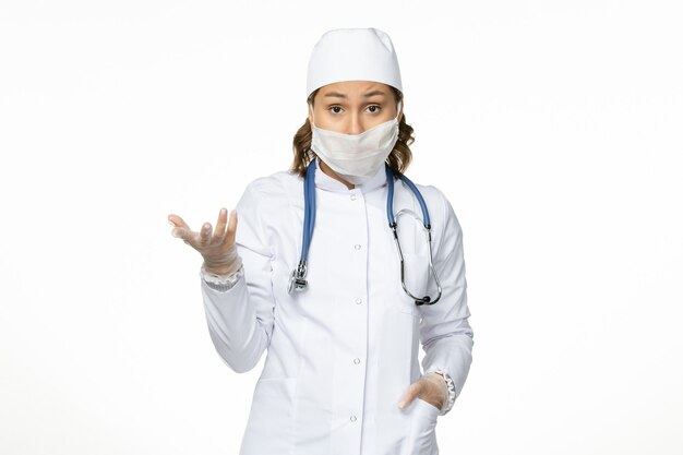 Vue de face jeune femme médecin en costume médical blanc et avec masque en raison d'un coronavirus sur surface blanche