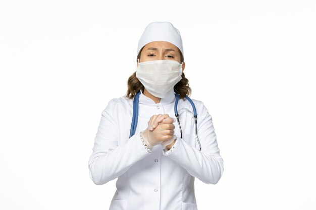 Vue de face jeune femme médecin en costume médical blanc et avec masque stérile en raison d'un coronavirus sur une surface blanche