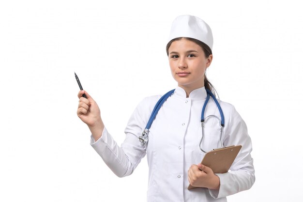 Une vue de face jeune femme médecin en costume médical blanc avec stéthoscope bleu écrit des notes