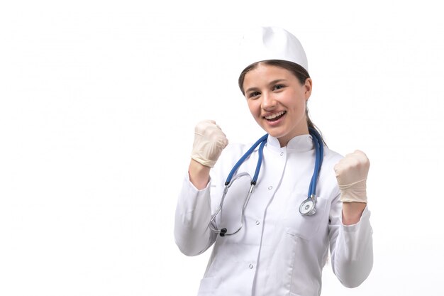 Une vue de face jeune femme médecin en costume médical blanc avec stéthoscope bleu portant des gants heureux