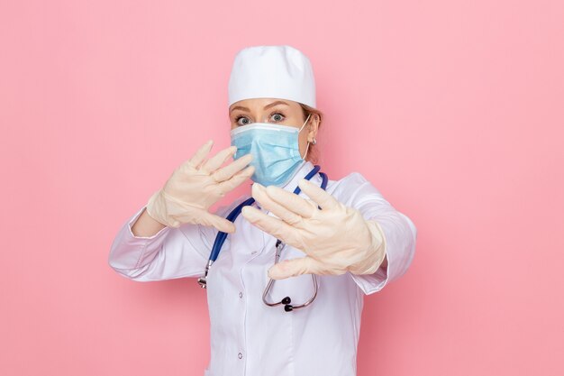 Vue de face jeune femme médecin en costume médical blanc avec stéthoscope bleu portant un masque stérile posant sur l'espace rose