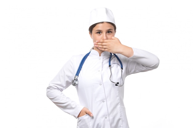 Une vue de face jeune femme médecin en costume médical blanc avec stéthoscope bleu posant avec une expression timide