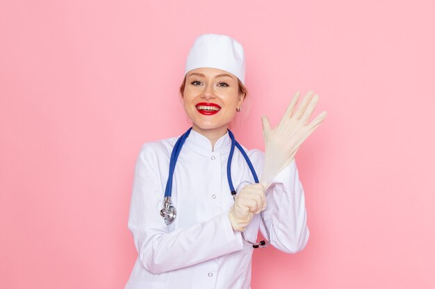 Vue de face jeune femme médecin en costume médical blanc avec stéthoscope bleu souriant et portant des gants sur l'hôpital médical de médecine de l'espace rose