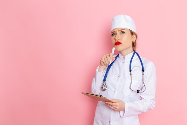 Vue de face jeune femme médecin en costume médical blanc avec stéthoscope bleu tenant le bloc-notes et en pensant à l'espace rose