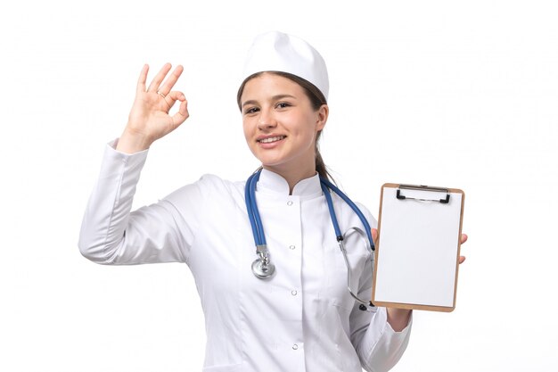 Une vue de face jeune femme médecin en costume médical blanc avec stéthoscope bleu tenant le bloc-notes