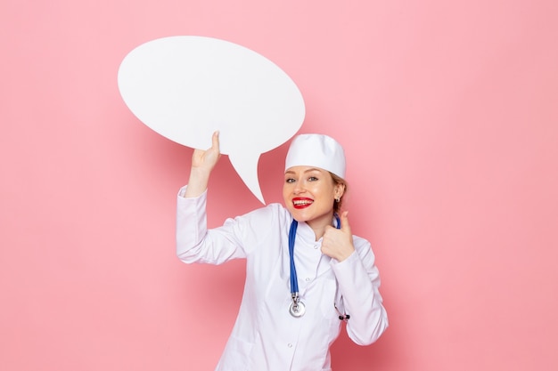 Vue de face jeune femme médecin en costume médical blanc avec stéthoscope bleu tenant un énorme panneau blanc avec sourire sur l'espace rose