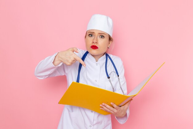 Vue de face jeune femme médecin en costume médical blanc avec stéthoscope bleu tenant des fichiers jaunes sur l'emploi de l'espace rose