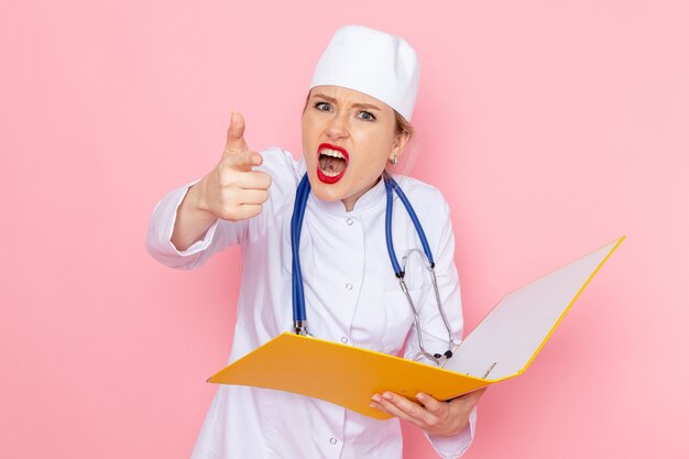 Vue de face jeune femme médecin en costume médical blanc avec stéthoscope bleu tenant des fichiers jaunes menaçant sur l'espace rose