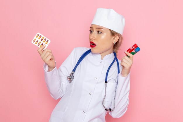 Vue de face jeune femme médecin en costume médical blanc avec stéthoscope bleu tenant des pilules et des flacons sur l'espace rose