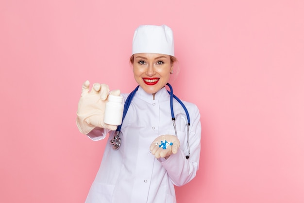 Vue de face jeune femme médecin en costume médical blanc avec stéthoscope bleu tenant des pilules avec sourire sur l'espace rose