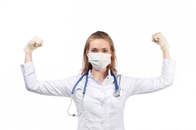 Une vue de face jeune femme médecin en costume médical blanc avec stéthoscope portant un masque de protection blanc dans des gants fléchissant sur le blanc