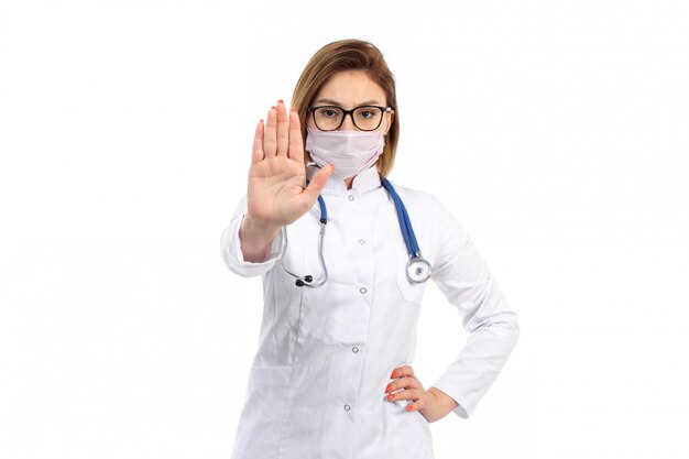 Une vue de face jeune femme médecin en costume médical blanc avec stéthoscope portant un masque de protection blanc montrant panneau d'arrêt sur le blanc