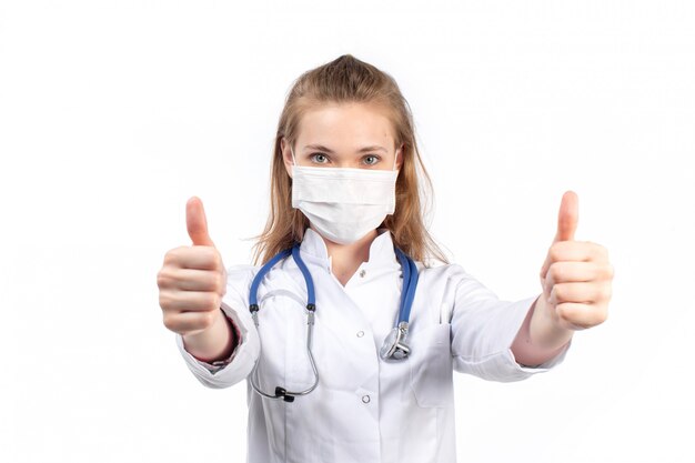 Une vue de face jeune femme médecin en costume médical blanc avec stéthoscope portant un masque de protection blanc posant montrant comme signe sur le blanc