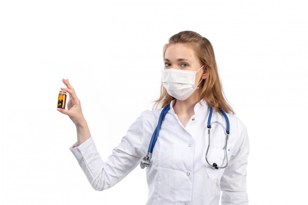 Une vue de face jeune femme médecin en costume médical blanc avec stéthoscope portant un masque de protection blanc posant tenant des pilules sur le blanc