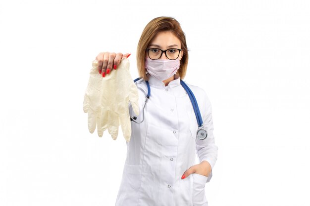 Une vue de face jeune femme médecin en costume médical blanc avec stéthoscope portant un masque de protection blanc tenant des gants sur le blanc