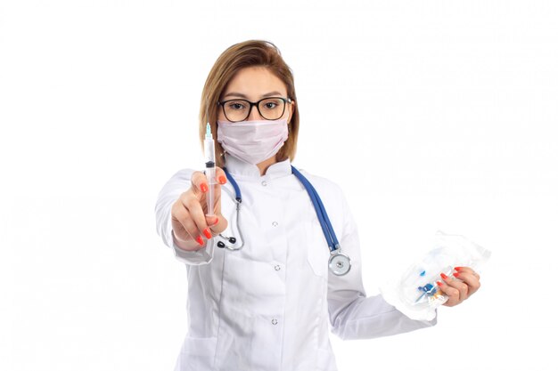 Une vue de face jeune femme médecin en costume médical blanc avec stéthoscope portant un masque de protection blanc tenant l'injection sur le blanc