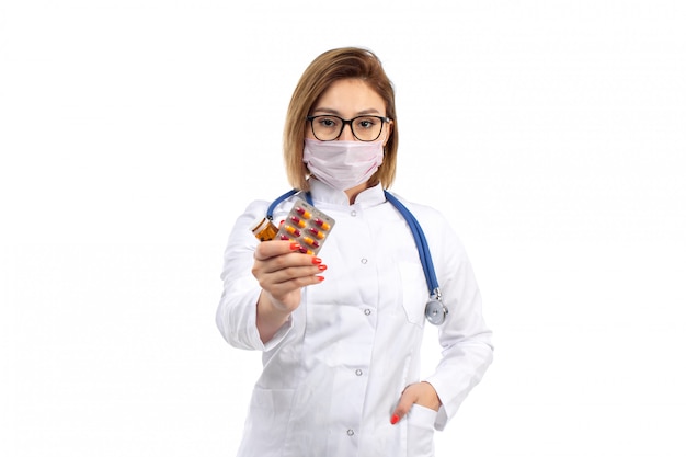 Une vue de face jeune femme médecin en costume médical blanc avec stéthoscope portant un masque de protection blanc tenant des pilules sur le blanc