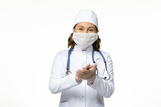 Vue de face jeune femme médecin avec masque de protection en raison du coronavirus et souriant sur une surface blanche