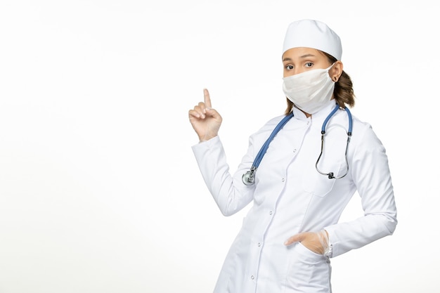 Vue de face jeune femme médecin avec masque stérile en raison d'un coronavirus sur la surface blanche claire