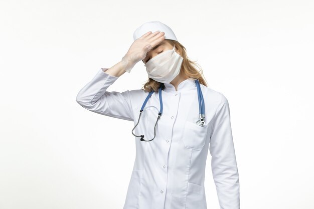 Vue de face jeune femme médecin portant un masque stérile de protection en raison d'un coronavirus sur une surface blanche