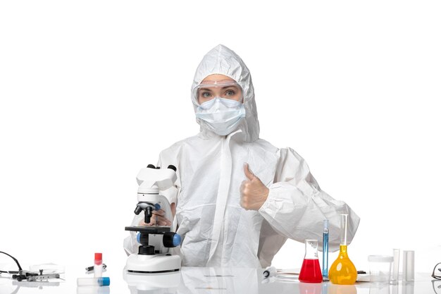 Vue de face jeune femme médecin en tenue de protection blanche avec masque en raison de covid à l'aide d'un microscope sur fond blanc clair santé éclaboussure pandémique covid-