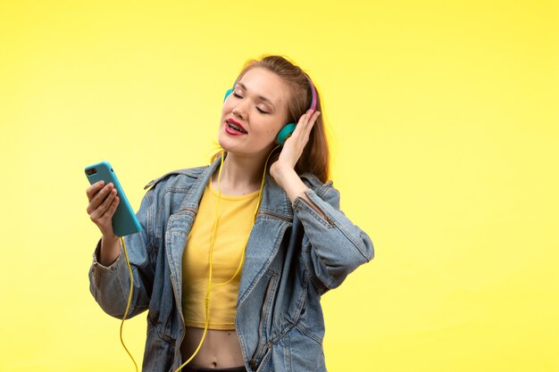 Une vue de face jeune femme moderne en chemise jaune pantalon noir et manteau en jean avec des écouteurs colorés écouter de la musique posant